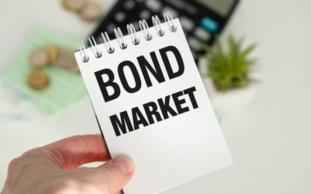 A person is holding a notebook with the word Bond Market written on it. The notebook is on a table with a calculator and some coinsの写真素材