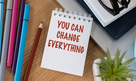 A notebook with the words You Can Change Everything written on it. The notebook is sitting on a desk with a pencil, a pen, and a pair of glassesの写真素材