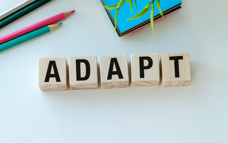 A wooden block with the word Adapt written on it. The block is placed on a white surface, and there are several colorful pencils and a potted plant nearby. Concept of creativity and learningの写真素材