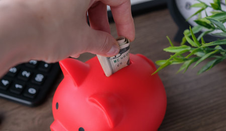 Close-up of a person saving money by putting a US dollar banknote into a red piggy bank. Business...