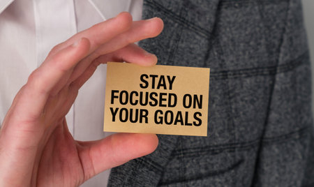 A close-up shot of a hand holding a small yellow card with the inspirational phrase STAY FOCUSED ON YOUR GOALS in bold black letters. The background shows a person wearing a professional gray suit jacket and a white shirt, symbolizing business leadership, personal growth, and determination.