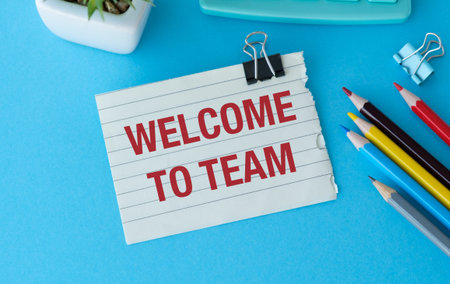 Blue desk holds a lined note card. Red text reads WELCOME TO TEAM. Black clips secure the note in place. Colored pencils lie nearby, ready for use. Light blue background adds calm, professional tone