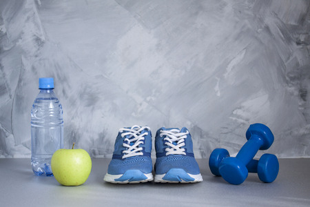 Sport shoes, dumbbells, apple, bottle of water on gray concrete background. Concept healthy lifestyle, sport and diet. Sport equipment.の写真素材