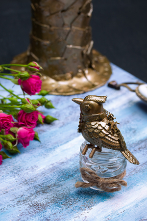 Steampunk bronze bird on glass cup, pink flowers, metallic hat on blue wooden tray.の写真素材