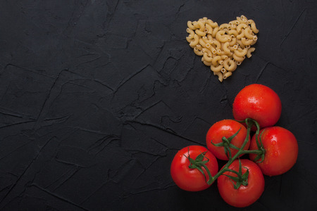 Red fresh large organic tomatoes with green leaves and pasta lying in shape of heart on black concrete background. Horizontal orientation, top view, place for copyspace.の写真素材