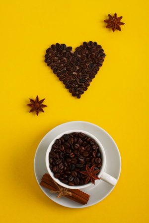 Cup of coffee seeds, cinnamon, anis and coffee heart shape on yellow background. I love coffee, coffee love concept. Flat lay, close up.の写真素材