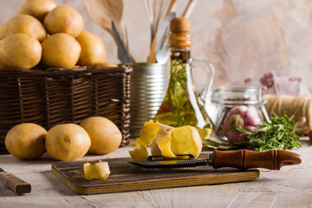 Peeling potatoes on dark cutting board on kitchen background. Wooden basket with potatoes, knife, bottle of oil, rosemary, garlic on beige background.の写真素材