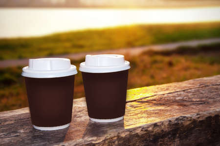 two paper cups with a lid for coffee to take away. Takeaway tea stands on a bench by the riverbankの写真素材