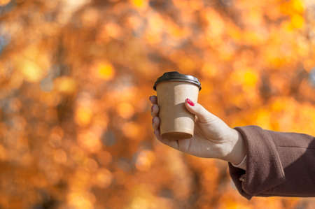 paper glass with coffee or tea in the girls hand against the background of autumn leaves, blur,...
