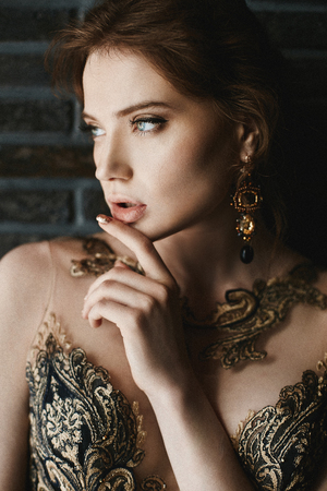 Beautiful, sexy, fashionable brunette girl with blue eyes, in gold dress with handmade gold earrings, is posing at brick wall background.の写真素材