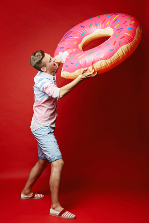 Young fashionable blond men in a shirt and shorts with inflatable donut, isolated at red background.の写真素材