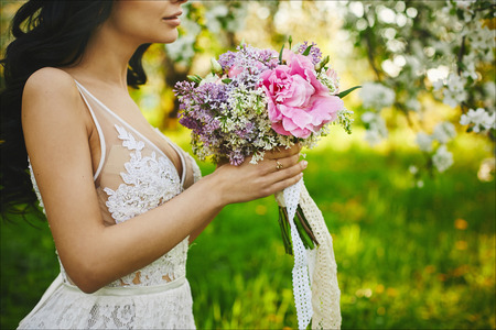Beautiful young bride, model brunette girl in white dress, with flowers in her hands, posing outdoors.の写真素材