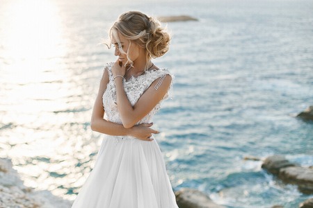 Beautiful young blonde model girl, in white lace dress, stands over the sea coastの写真素材