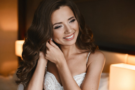 Portrait of happy, beautiful and fashionable brunette model girl with professional bright makeup and hairstyle, in lace dress posing and adjusting earring, wedding preparation of young brideの写真素材
