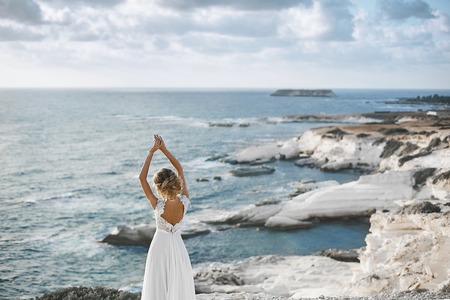 Beautiful young blonde model girl, in white lace dress with naked back, stands with raised hands at the coast and looks at the sea.の写真素材