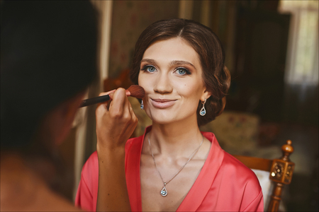 Makeup artist applies makeup for beautiful, smiling and cheerful young brunette woman with blue eyes and luxury earrings in dressing gown, wedding preparation of pretty brideの写真素材