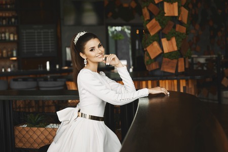 Young elegant beautiful and sexy brunette model woman in white dress with gold belt sitting in bar and waiting for boyfriend.の写真素材