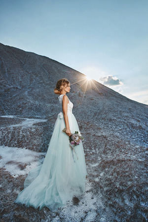 Fashionable and beautiful blonde model girl with modeling hairstyle in stylish white lace dress with bouquet of exotic flowers in her hand posing on the mountain at the sunsetの写真素材