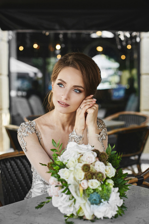 Portrait of beautiful and sensual brunette model girl with bright makeup and blue eyes in fashionable lace dress sitting at a table with bouquet of flowers in a cafe outdoorsの写真素材
