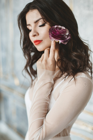 Portrait of sensual and beautiful brunette model girl with bright makeup and closed eyes in stylish light peignoir with flower in her hand posing in luxury vintage interiorの写真素材
