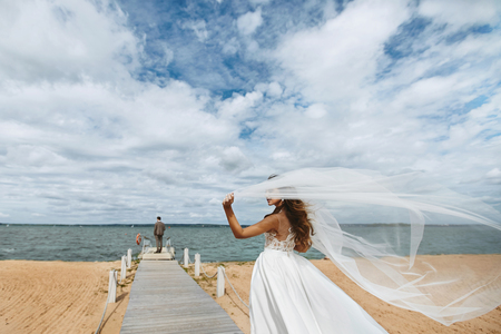Young bride, fashionable and beautiful brunette model girl in stylish wedding dress with a veil which fluttering in the wind, walks to the groom who waiting at the pierの写真素材