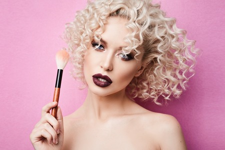 Fashionable and stylish model girl with blue eyes and curly blond hair, with professional bright makeup posing with makeup brush in her hand at studio, isolated at pink backgroundの写真素材