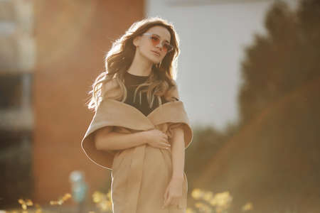 Fashionable beautiful and sensual blonde model girl in sleeveless coat and stylish sunglasses enjoys the sun and posing outdoors at the city street.の写真素材