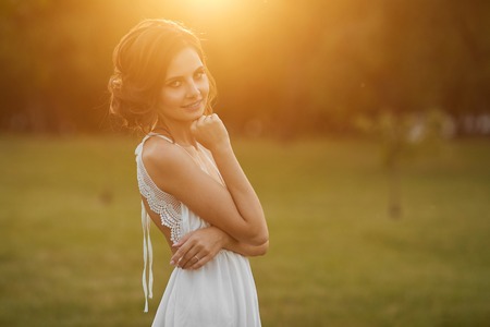 Beautiful and young brunette model girl with gentle makeup, in short white dress stands and posing at green park in early evening at the sunsetの写真素材