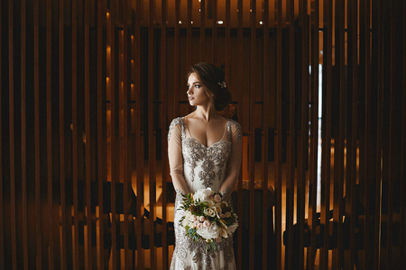 Beautiful and sensual brunette model girl with bright makeup and wedding hairstyle, in fashionable white lace dress with bouquet of flowers in her hands posing at luxury stylish interior.の写真素材