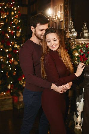 Fashionable and beautiful couple, stylish handsome bearded men hugs the sexy brunette model girl in a red fitting dress and posing near the Christmas tree at interior decorated for New Yearの写真素材
