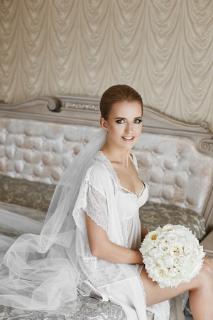 Sexy blonde model girl with blue eyes and gentle makeup, in lace peignoir and in white lingerie with the bouquet of peony flowers in her hands sit on the bed and posing at luxury interiorの写真素材