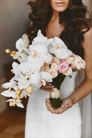 Fashionable and elegant brunette model girl with stylish wedding hairstyle and with bright makeup in white dress with the big luxury bouquet of exotic flowers in her hands posing at interiorの写真素材