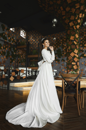 Elegant and beautiful brunette model girl with bright makeup and with stylish hairstyle with jewelry in the white fashionable long dress smiles and posing at luxury interior in a cafeの写真素材