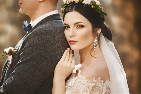 Elegant and fashionable bride, sensual young model woman in trendy lace dress with stylish hairstyle and with bright make-up lean on handsome man in suit and posing outdoorsの写真素材