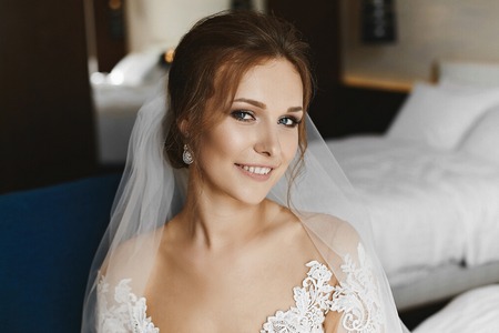 Beautiful and stylish brown-haired model girl with professional makeup and with big luxury earrings in lace wedding dress smiles and posing in interiorの写真素材