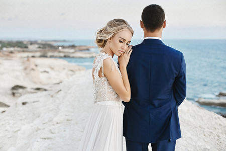 Beautiful young blonde model girl in fashionable white lace dress lean on the handsome man in the stylish blue suit and posing on the white rock at the coast of the Adriatic seaの写真素材