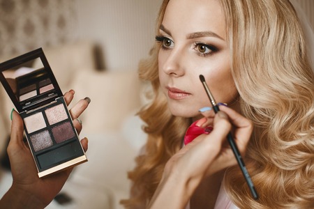 Visagiste applies makeup for a beautiful blonde girl with wedding hairstyleの写真素材
