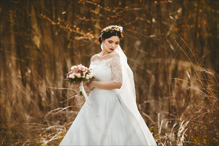 Elegant and fashionable young brunette bride with bright makeup and with a floral wreath on her head in the stylish lace wedding dress with a bouquet of flowers in her hands posing outdoorsの写真素材