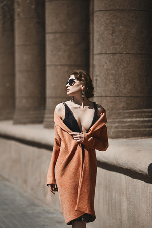 Beautiful brunette girl with sexy body in the black stylish dress, in a trendy coat and in fashionable sunglasses posing at the city street, live photoの写真素材