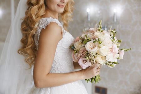 Beautiful young blonde bride with stylish wedding hairstyle in a white fashionable dress with a bouquet of flowers in her handsの写真素材