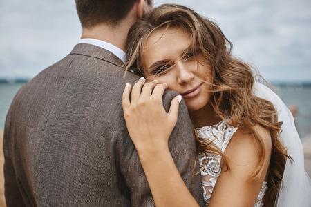 Elegant and happy model girl with stylish hairstyle and with bright makeup in a white lace dress lean on her stylish groomの写真素材