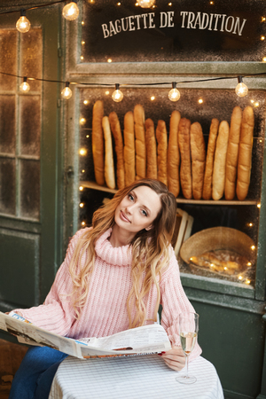 Fashionable and beautiful blonde model girl in trendy oversize sweater sit with newspaper and glass of champagneの写真素材