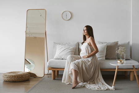 Beautiful brown-haired pregnant model girl with blue eyes and with a pretty smile in a stylish beige dress sits on the sofa and posing at minimalist interiorの写真素材