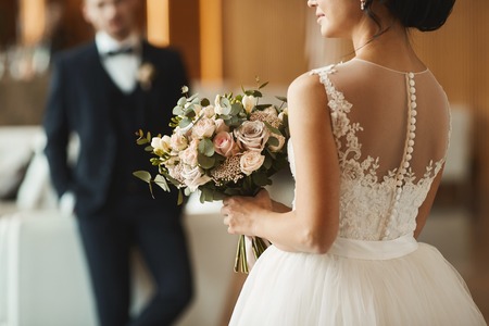 Fashionable couple, handsome young man in the stylish suit and beautiful brunette model girl with wedding hairstyle in white lace dress holding a bouquet of flowersの写真素材