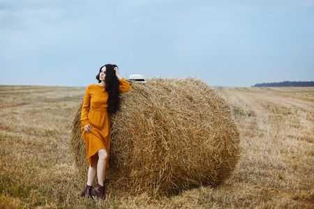 Beautiful brunette model girl in the fashionable dress of mustard color posing near the stack of hay at the fieldの写真素材