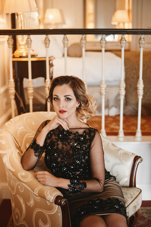 Young and sexy businesswoman in a stylish cocktail dress sits in a vintage armchair at luxury interiorの写真素材