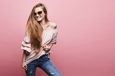 Happy and emotional young woman with blond long hair and with perfect body in stylish sunglasses and in the fashionable outfit at the pink background, isolatedの写真素材