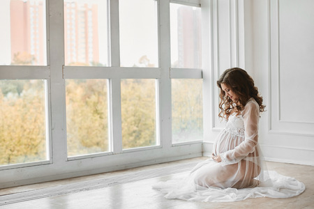 Beautiful young pregnant woman with fashionable hairstyle in a stylish peignoir smiling and posing near the big window at light interiorの写真素材