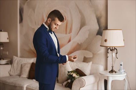 Handsome bearded young businessman in a stylish blue suit and in bowtie puts on stylish cufflinks and posing at interiorの写真素材