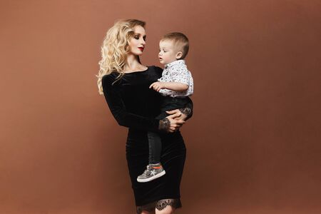 Beautiful and happy young blonde woman in a black dress with a cute little baby boy on her hands,...